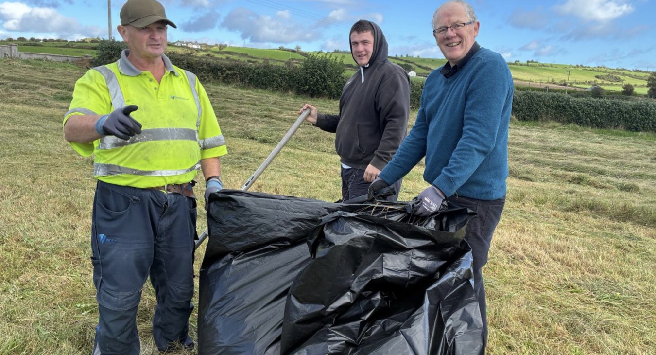 three men stand around a large black bag filled with green hay